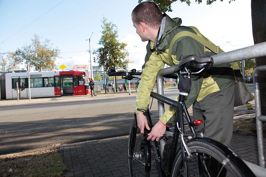 Öffentlicher Nahverkehr und Fahrrad Ein junger mann schließt sein Fahrrad an einem Fahrradbügel an und schaut zu einer Straßenbahn im Hintergrund.